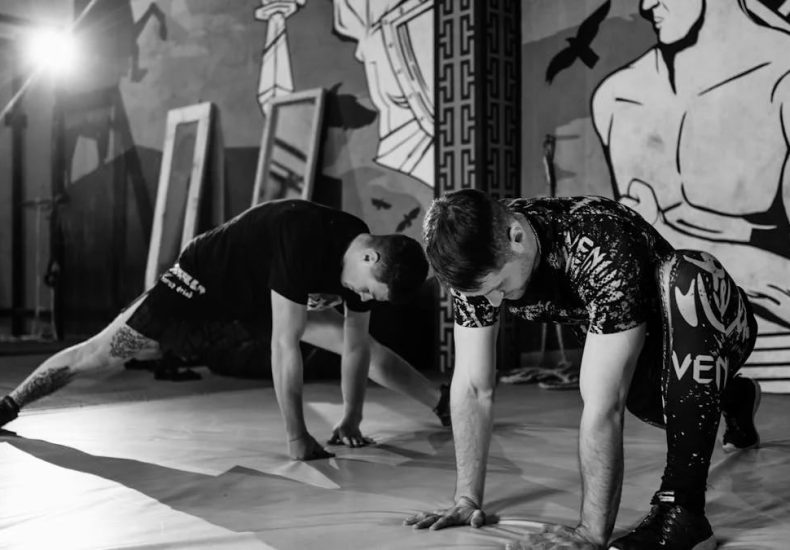 Two men stretching inside a gym, preparing for MMA training in a grayscale setting.